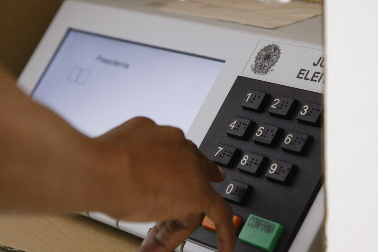 Mão de uma pessoa digitando na urna eletrônica; a tela mostra o cargo de Presidente.